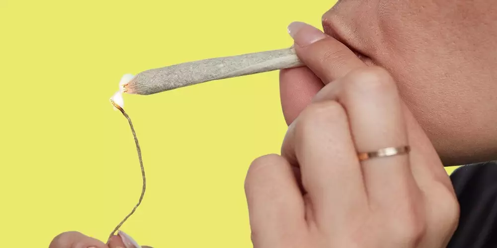 A woman is using a hemp wick to smoke marijuana on a yellow background.